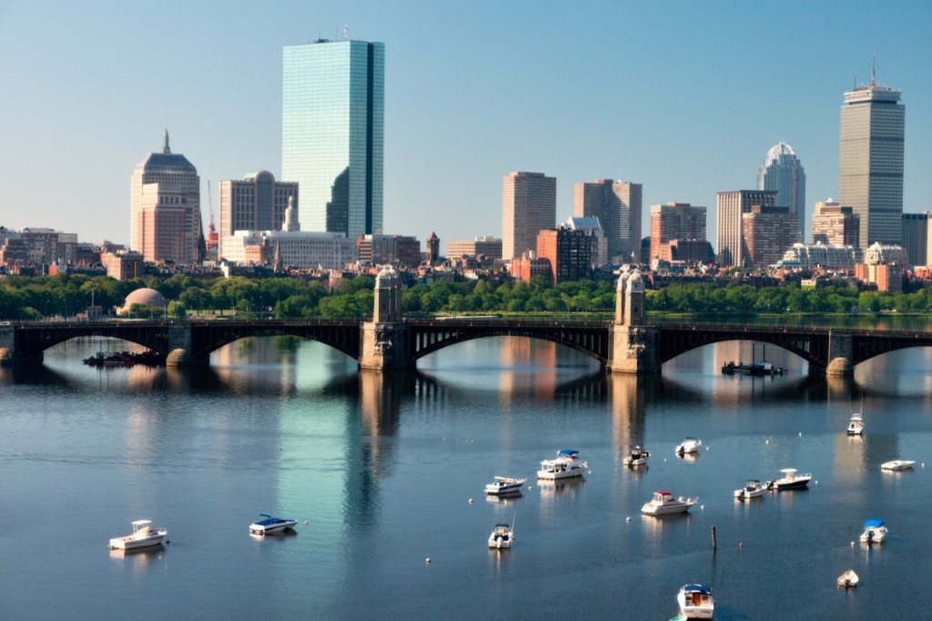 A view of a city skyline in Boston with modern buildings, including a tall glass structure, set against a clear blue sky. In the foreground, boats are anchored on a calm river with a bridge connecting the two sides.