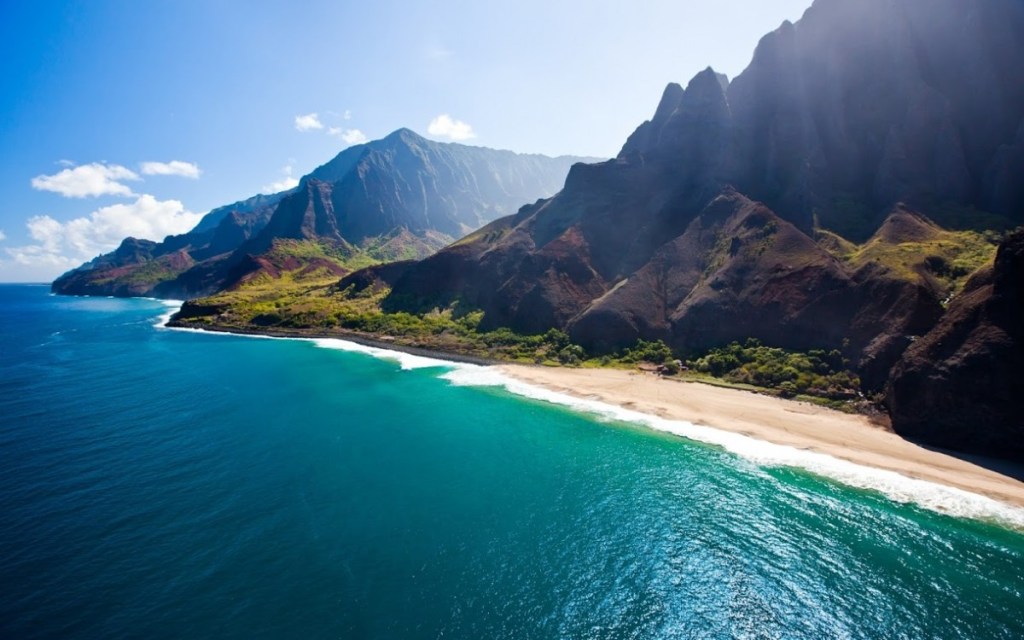 Scenic view of a rugged coastline in Kauai, Hawaii with mountains and a beach under a bright blue sky.